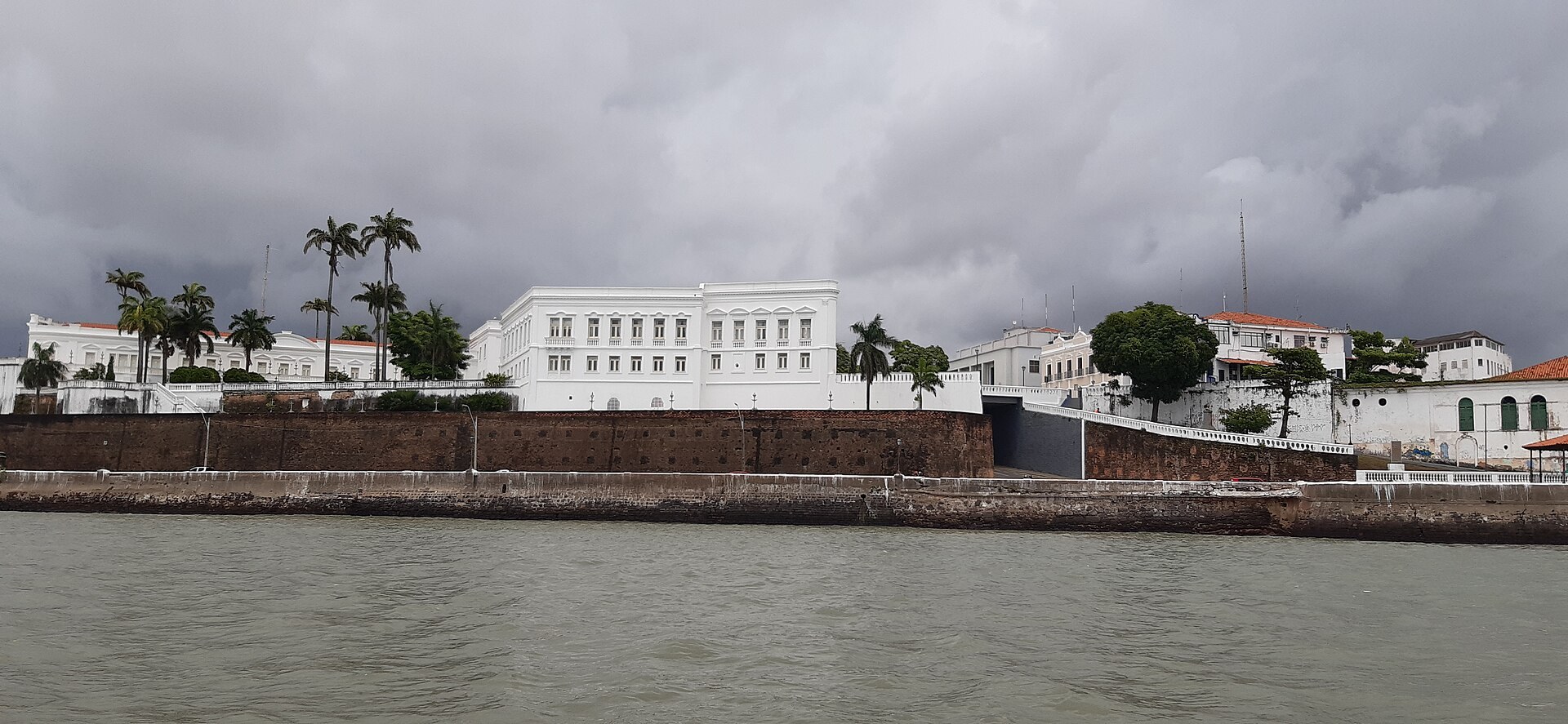 Palácio dos Leões em São Luís visto da água, muralha e palmeiras sob céu nublado.